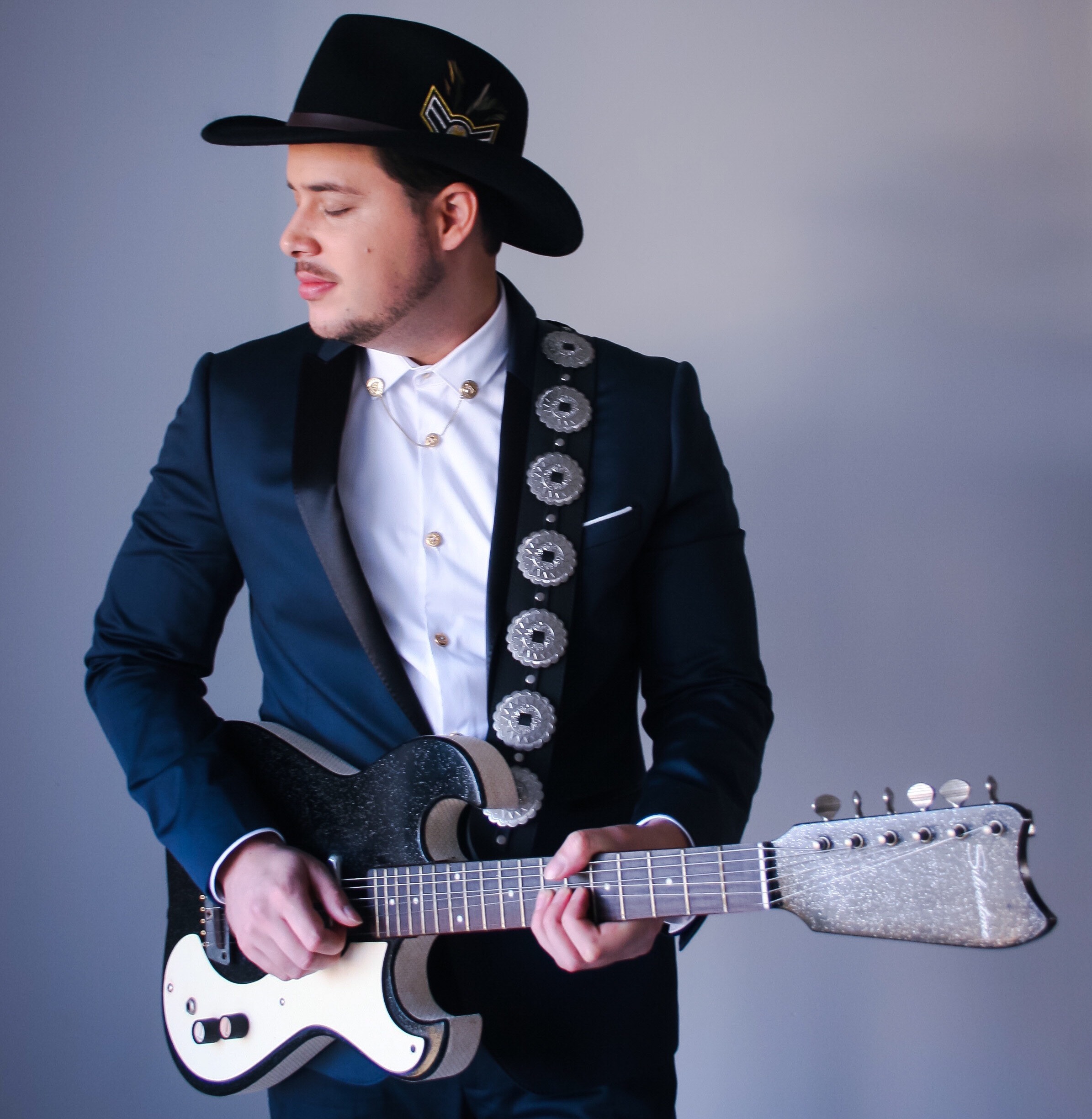 Formal studio portrait of Will Plus in a dark suit and hat holding an electric guitar