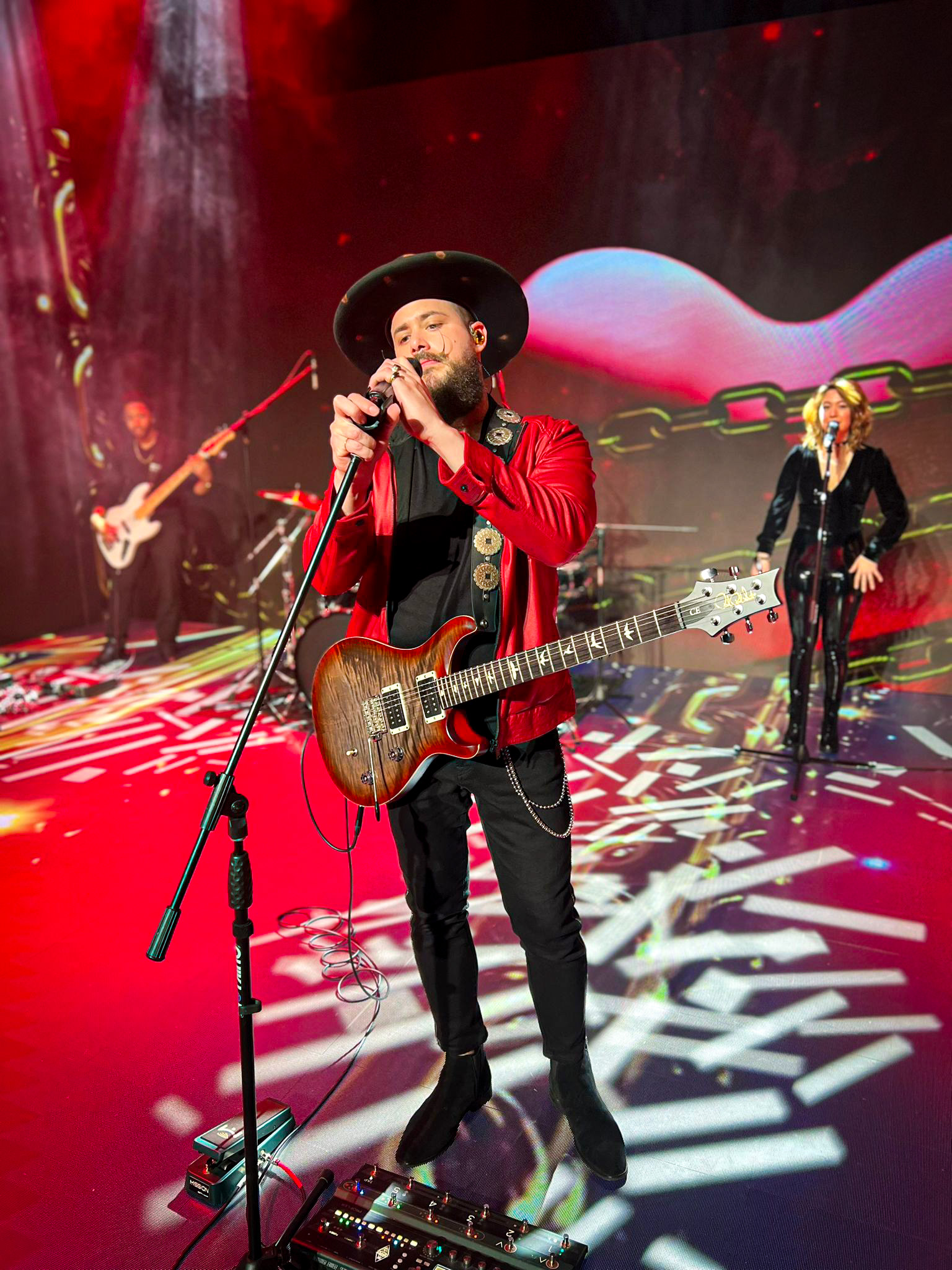 Will Plus singing with an electric guitar and live band on a red-lit concert stage